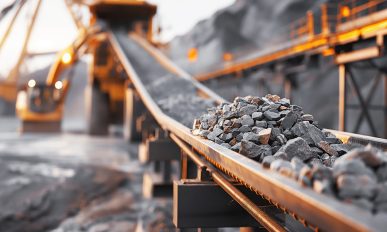 Close-up of rocks on a conveyor belt system in an industrial mining or quarry environment with heavy machinery in the background.